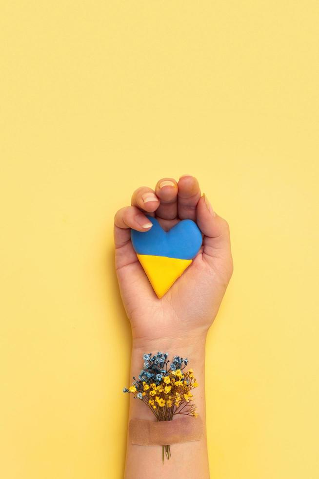 corazón y flor azul y amarilla debajo del parche en una mano femenina sobre un fondo amarillo. amar y orar al concepto de ucrania foto