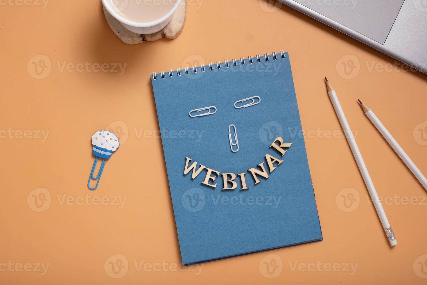 Webinar text from wooden letters on notepad on desktop with laptop and coffee on colored background. Top view, flat lay photo