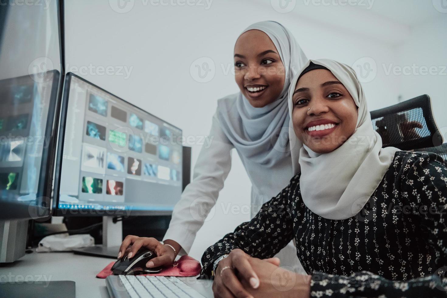 Friends at the office two young Afro American modern Muslim businesswomen wearing scarf in creative bright office workplace with a big screen photo