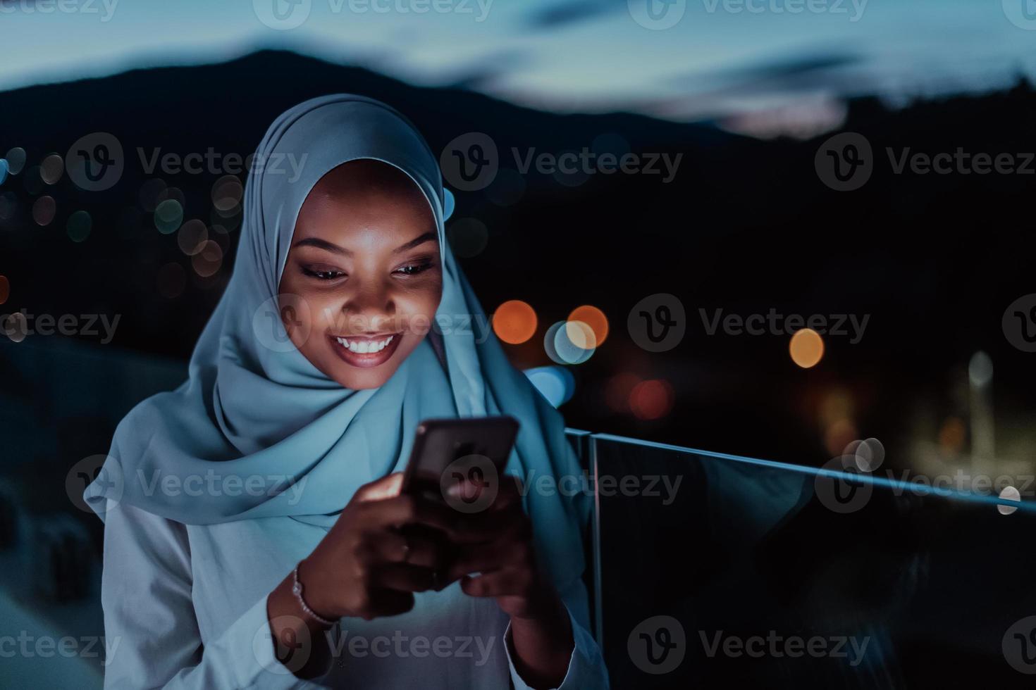 joven mujer musulmana con velo de bufanda en la calle urbana de la ciudad por la noche enviando mensajes de texto en un teléfono inteligente con la luz de la ciudad en el fondo. foto