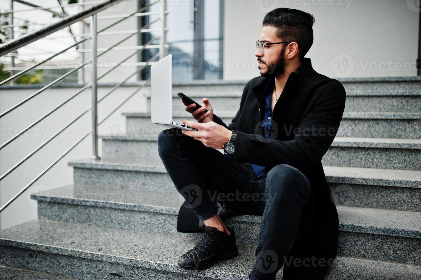 Middle eastern entrepreneur wear black coat and blue shirt, eyeglasses against office building with laptop. photo