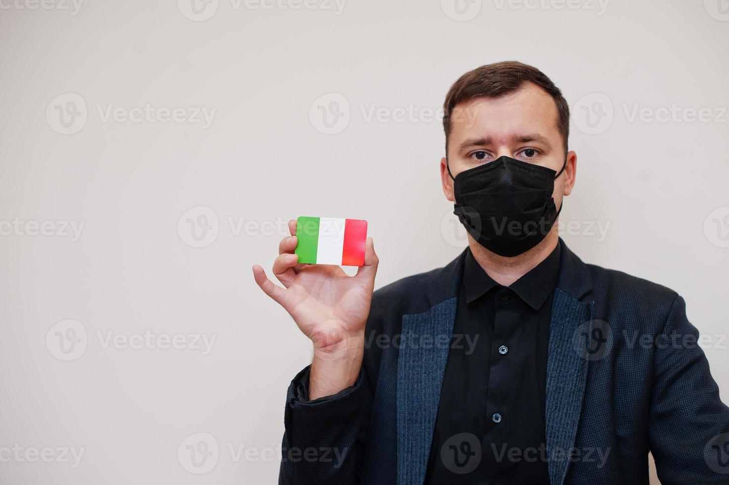 European man wear black formal and protect face mask, hold Italy flag card isolated on white background. Europe coronavirus Covid country concept. photo