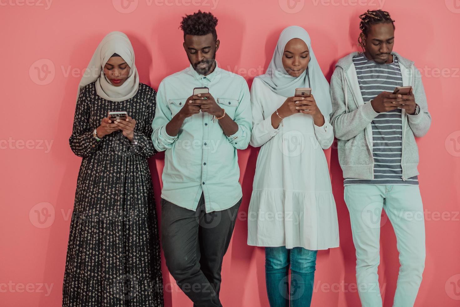 A group of African Muslim students use smartphones while standing in front of a pink background photo