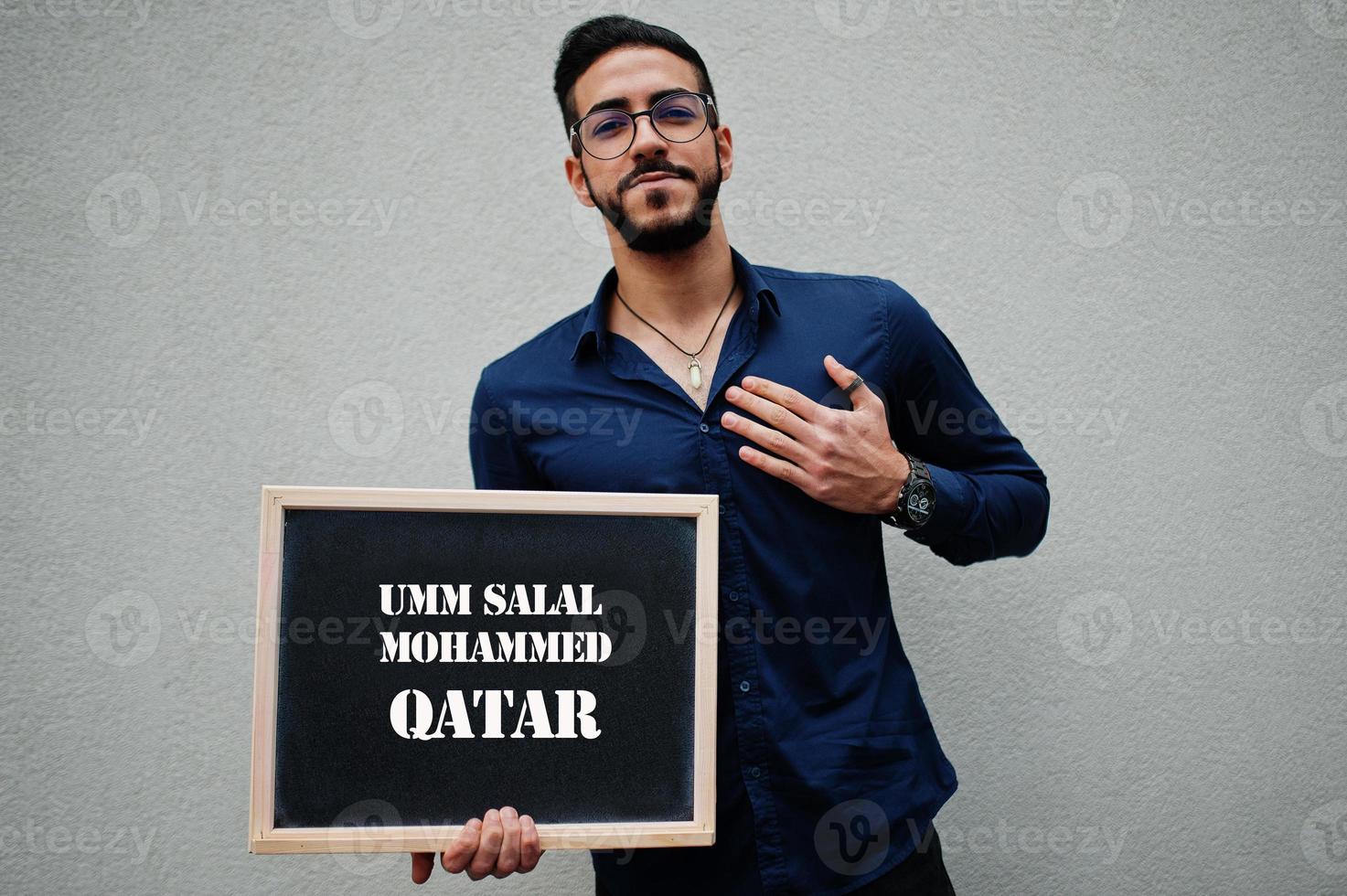 Arab man wear blue shirt and eyeglasses hold board with Umm Salal Mohammed Qatar inscription. Largest cities in islamic world concept. photo