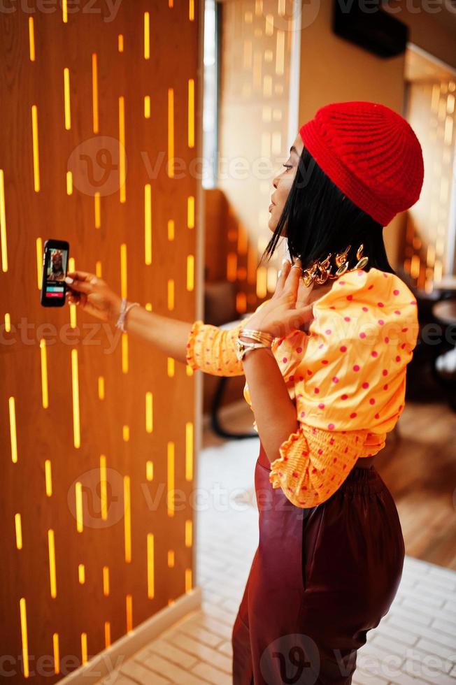 Elegant afro american woman in red french beret, big gold neck chain polka dot blouse and leather pants conducts a live broadcast by mobile phone for social network. photo