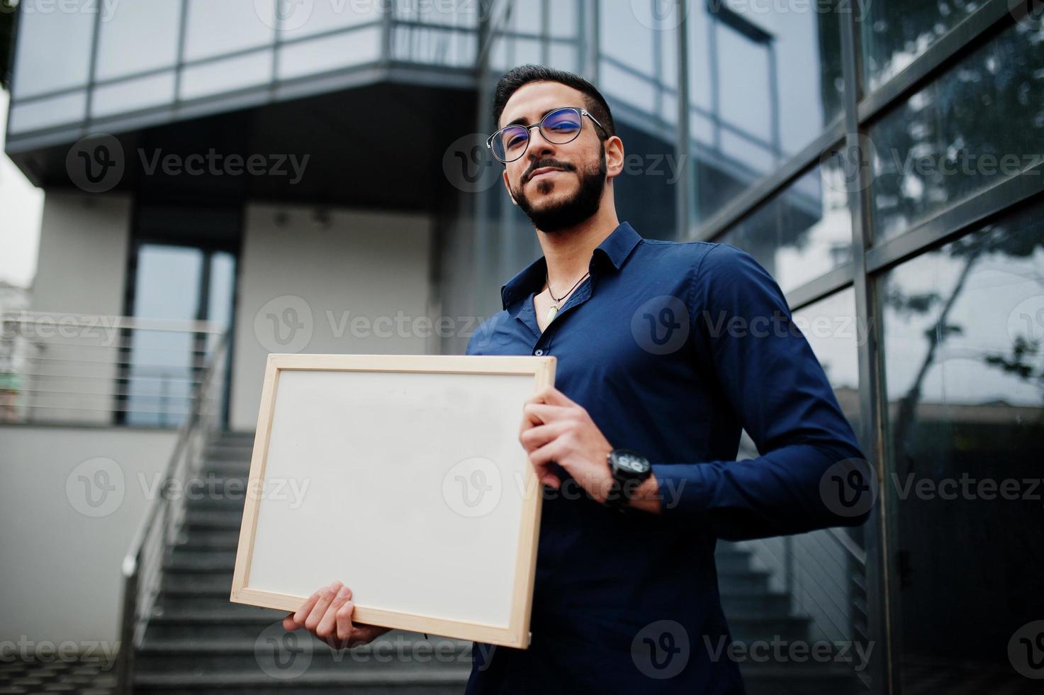 el hombre del medio oriente usa camisa azul, anteojos, sostiene un tablero blanco vacío. foto