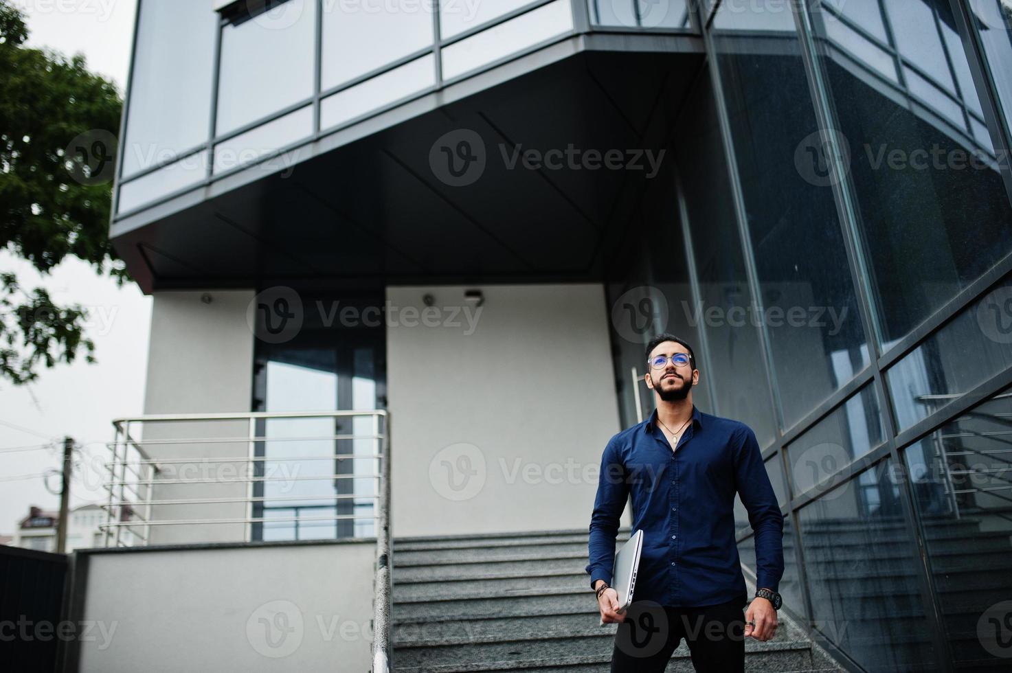empresario del medio oriente usa camisa azul, anteojos con computadora portátil contra el edificio de oficinas. foto