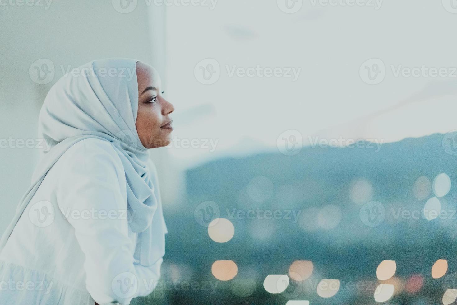 African Muslim woman in the night on a balcony smiling at the camera with city bokeh lights in the background. photo
