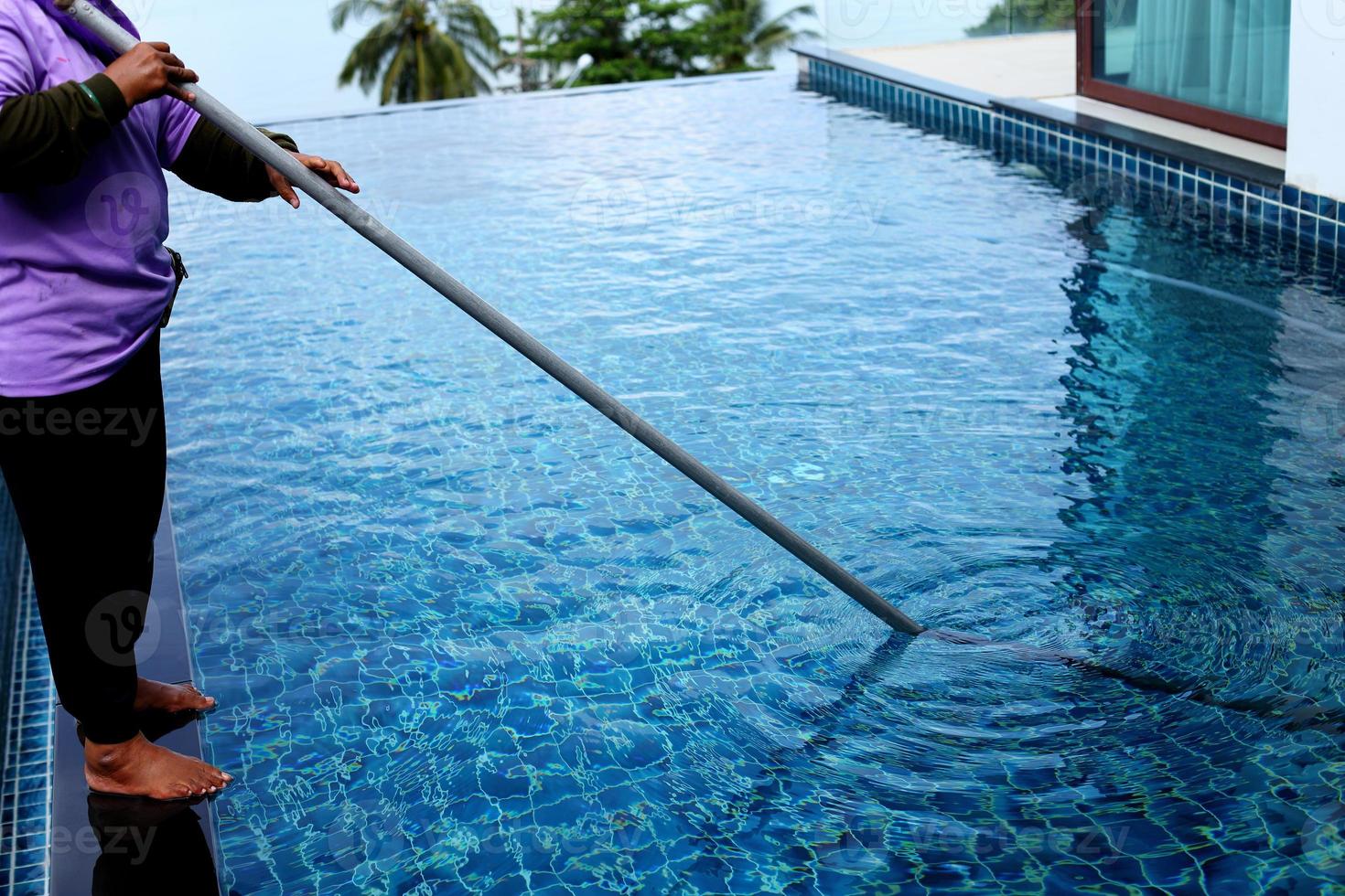 mujer trabajadora limpia piscina privada con limpiador de tubo azul al vacío cada semana en verano, piscina tipo sal junto con el océano como estilo horizontal con cocoteros foto