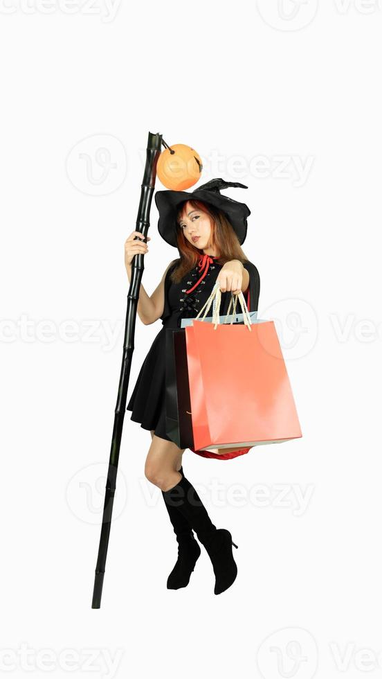 Beautiful girl with black and orange hair  in black dress and witch hat is holding staff with shopping bags on white background for Halloween day. photo