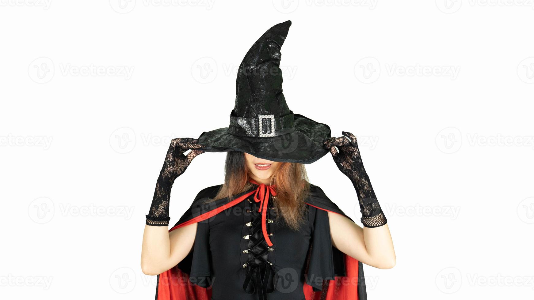 Gothic young woman in witch halloween costume with hat standing over white background. Beautiful young girl in black dress, covering her eyes with the cap photo