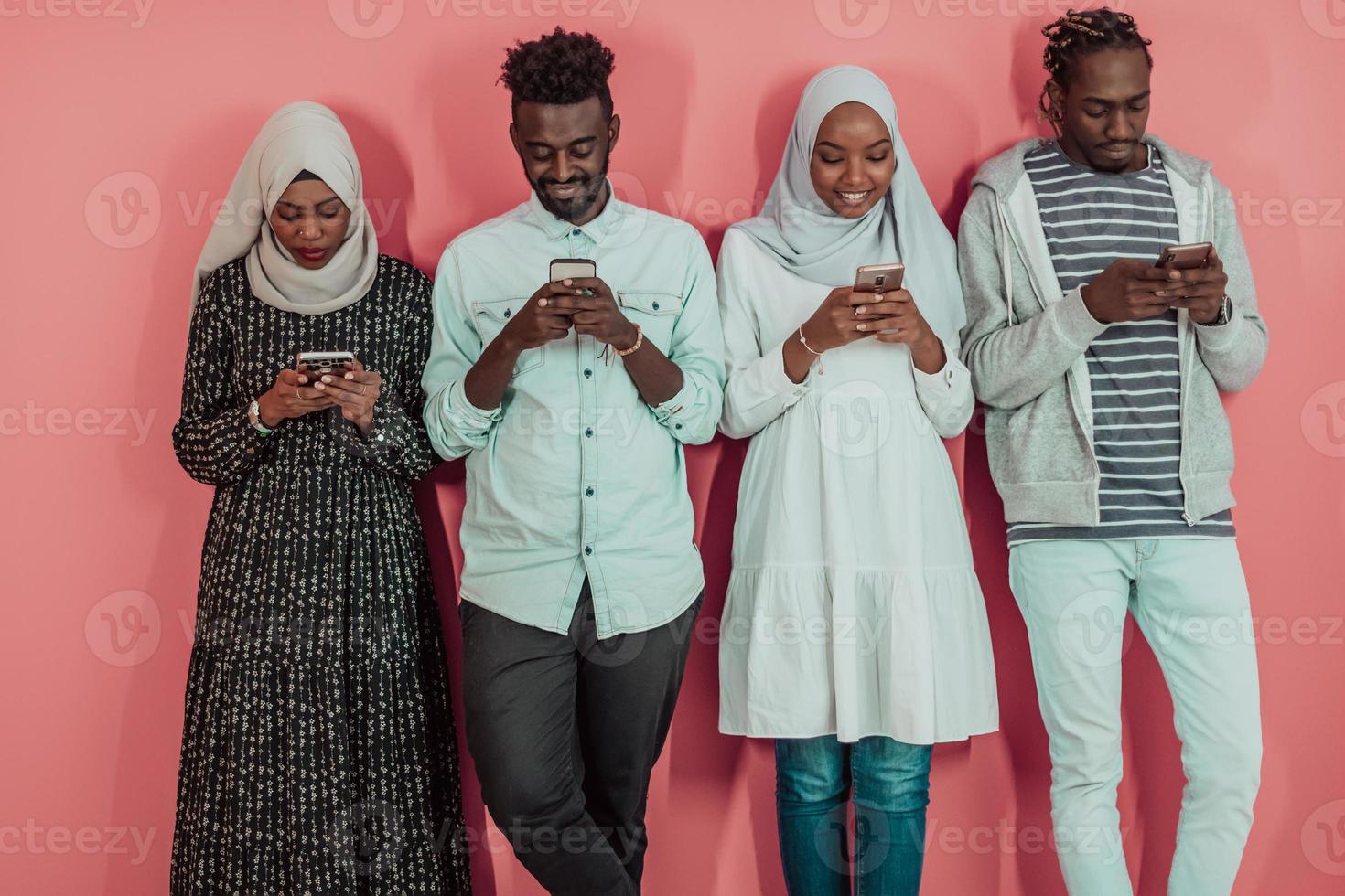 A group of African Muslim students use smartphones while standing in front of a pink background photo