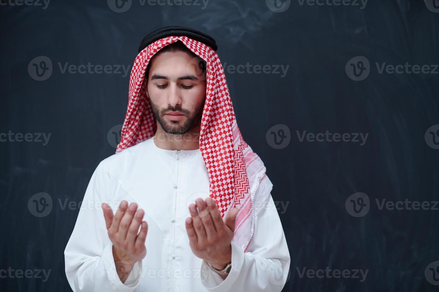 arabian man making traditional prayer to God, keeps hands in praying gesture in front of black chalkboard photo