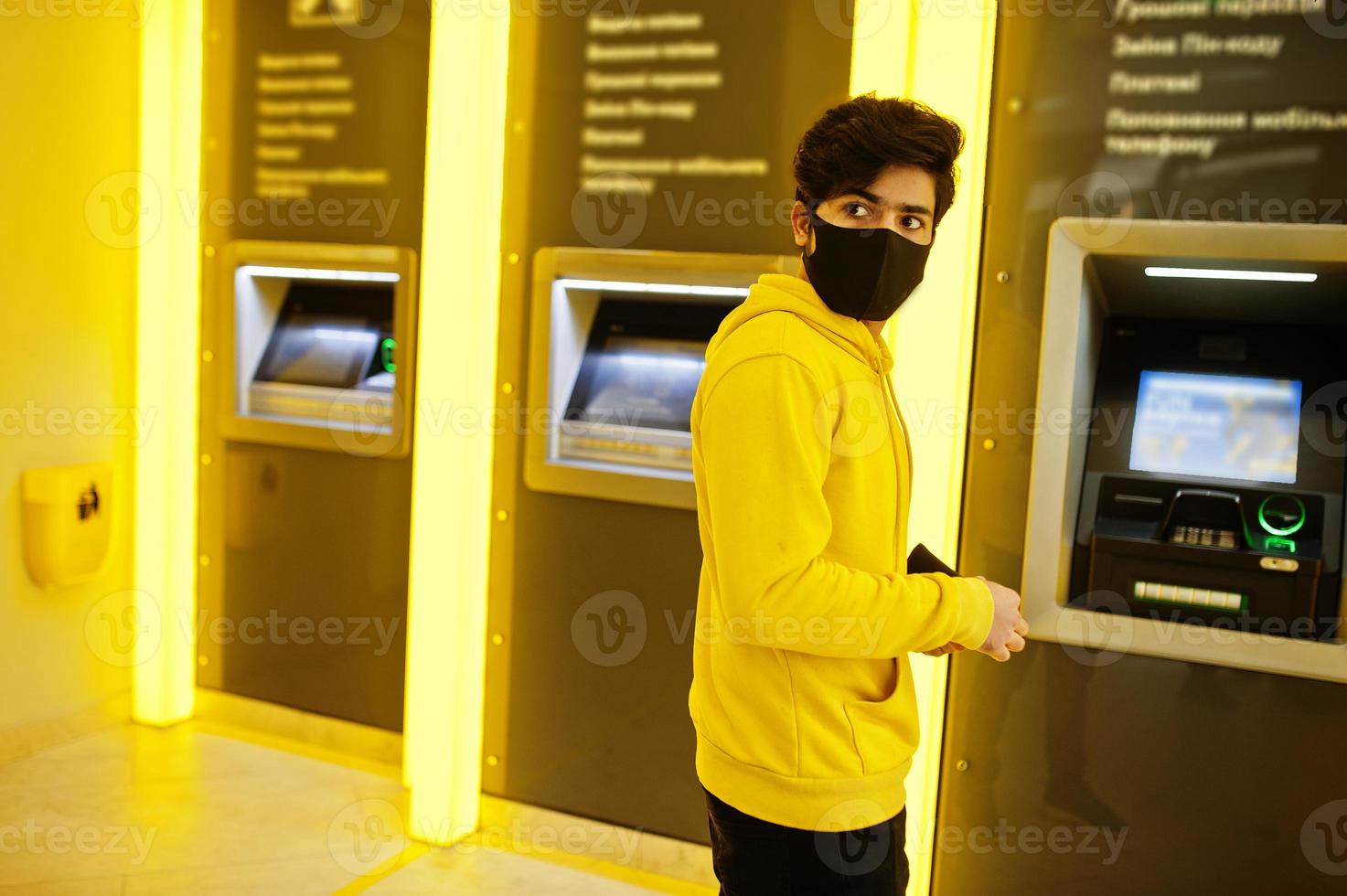 hombre indio joven hipster urbano en una sudadera amarilla de moda en el banco. un chico genial del sur de Asia usa una sudadera con capucha y una máscara de protección facial negra, durante la nueva normalidad, retira efectivo en el cajero automático. foto