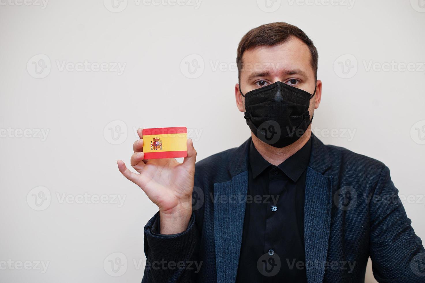 European man wear black formal and protect face mask, hold Spain flag card isolated on white background. Europe coronavirus Covid country concept. photo