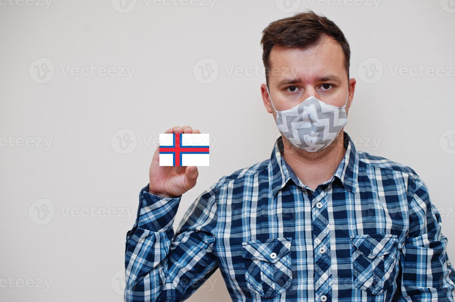 Man in checkered shirt show Faroe Islands flag card in hand, wear protect mask isolated on white background. Europe countries Coronavirus concept. photo