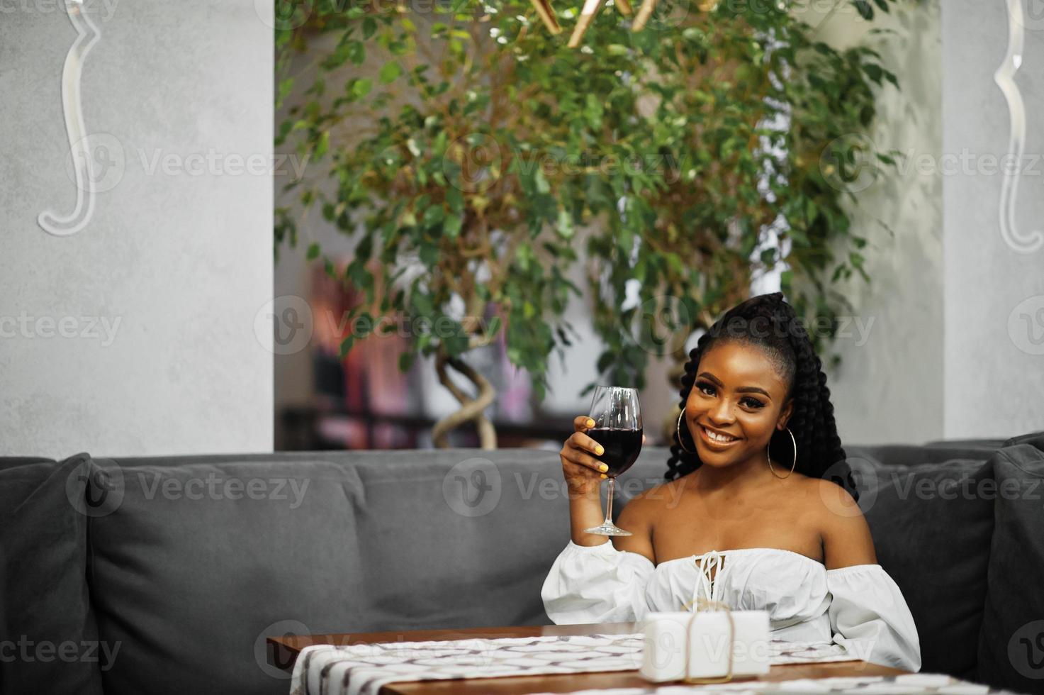 bella mujer afroamericana con blusa blanca y pantalones de cuero rojo posan en el restaurante con una copa de vino. foto