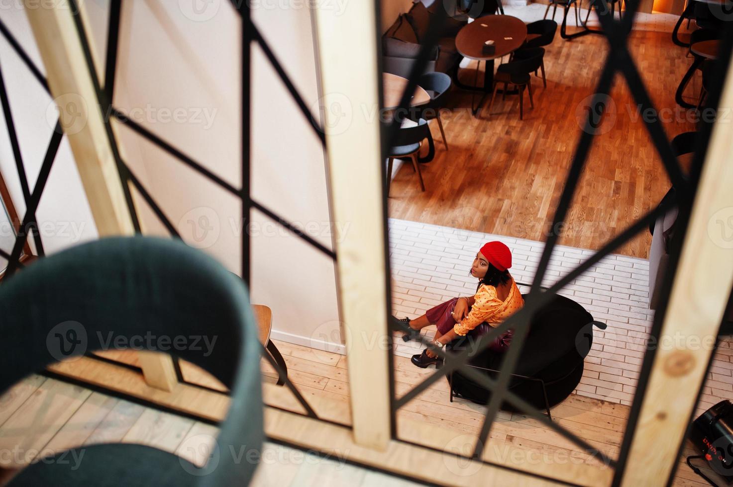 Elegant afro american woman in red french beret, big gold neck chain polka dot blouse and leather pants sit on pouf. photo