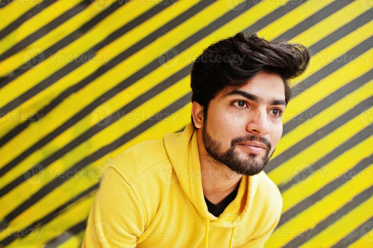 Urban young hipster indian man in a fashionable yellow sweatshirt. Cool south asian guy wear hoodie against striped background. photo