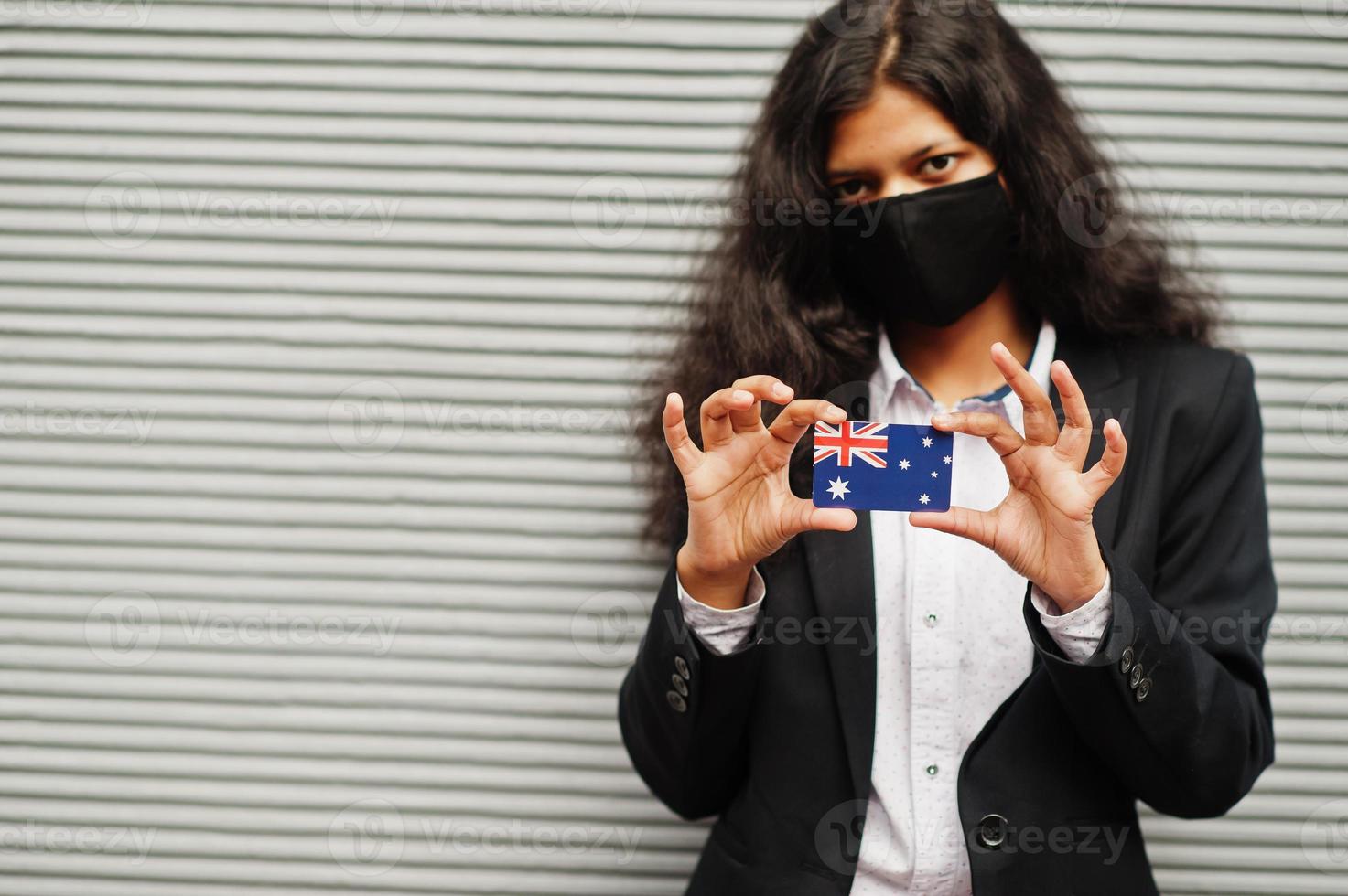 mujer asiática con ropa formal y máscara protectora negra sostiene la bandera de australia a mano contra un fondo gris. coronavirus en el concepto de país. foto