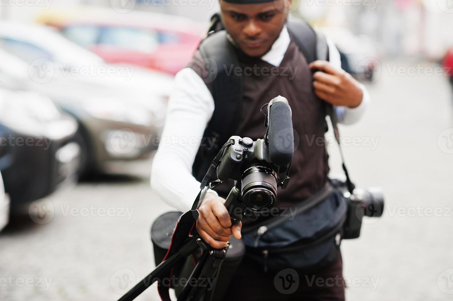 Young professional african american videographer holding professional camera with tripod pro equipment. Afro cameraman wearing black duraq making a videos. photo