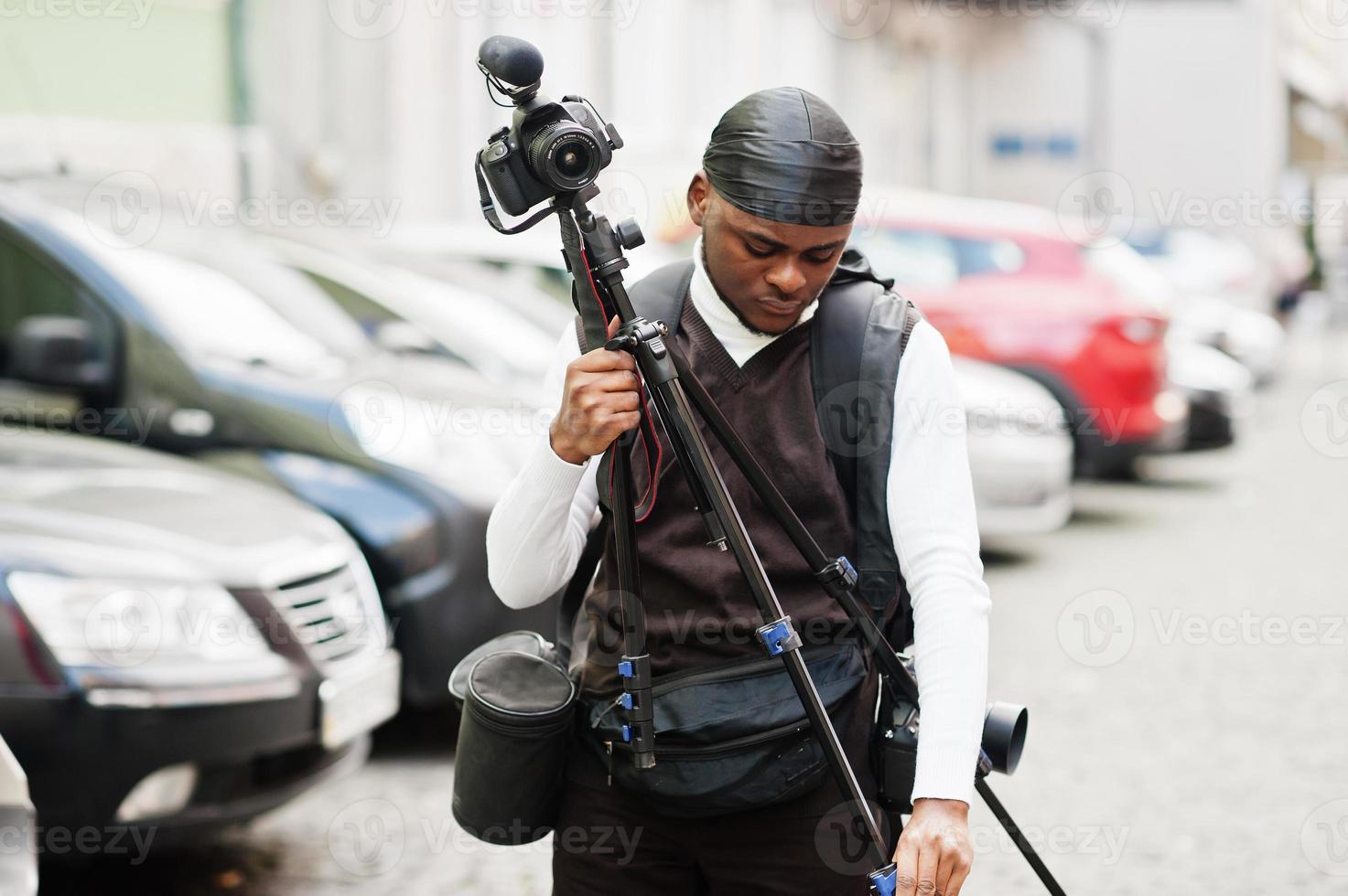 Young professional african american videographer holding professional camera with tripod pro equipment. Afro cameraman wearing black duraq making a videos. photo