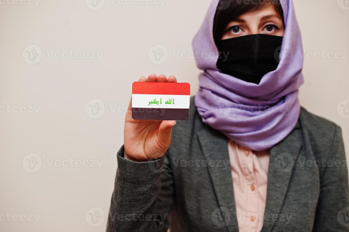 Portrait of young muslim woman wearing formal wear, protect face mask and hijab head scarf, hold Iraq flag card against isolated background. Coronavirus country concept. photo