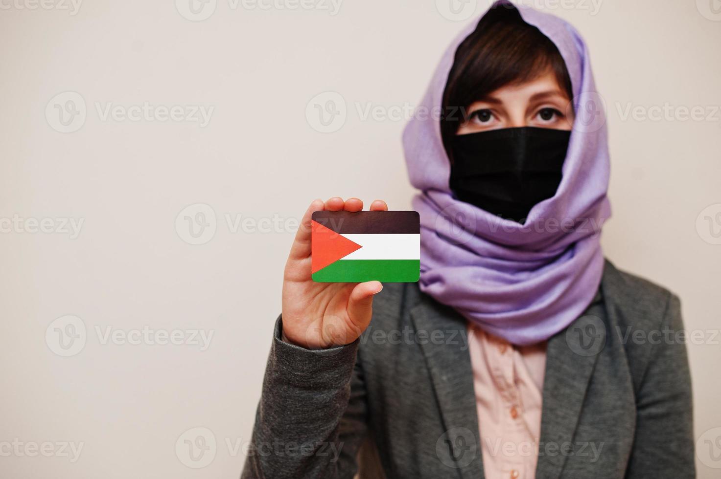 Portrait of young muslim woman wearing formal wear, protect face mask and hijab head scarf, hold Palestine flag card against isolated background. Coronavirus country concept. photo