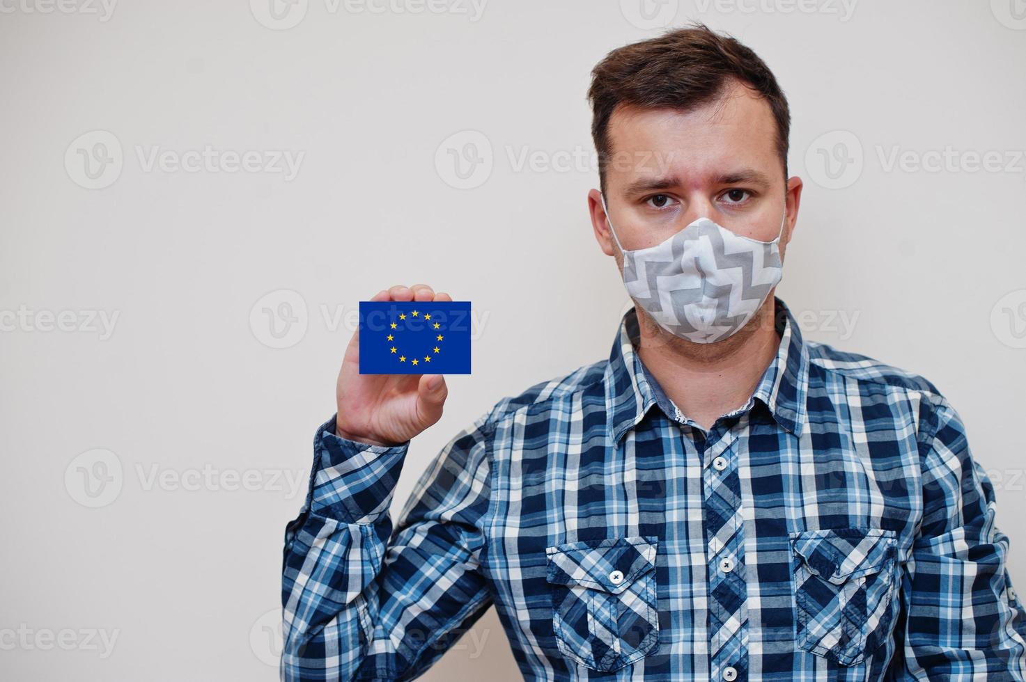 Man in checkered shirt show European Union flag card in hand, wear protect mask isolated on white background. Europe countries Coronavirus concept. photo