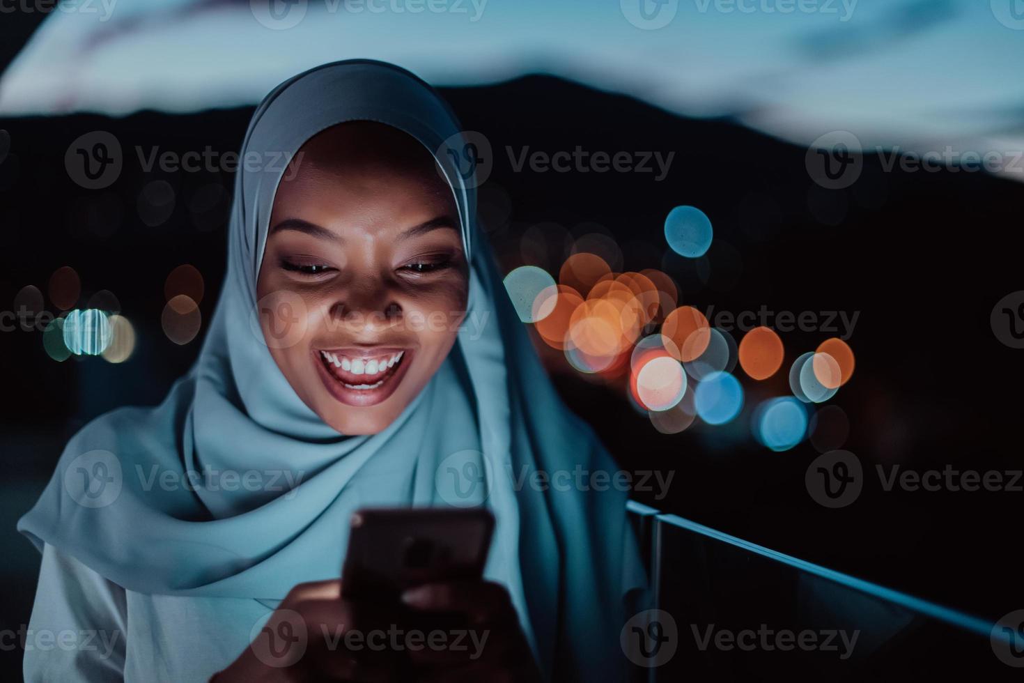 joven mujer musulmana con velo de bufanda en la calle urbana de la ciudad por la noche enviando mensajes de texto en un teléfono inteligente con la luz de la ciudad en el fondo. foto