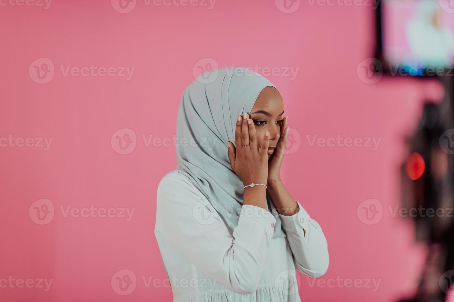 Modern African Muslim woman makes traditional prayer to God, keeps hands in praying gesture, wears traditional white clothes, has serious facial expression, isolated over plastic pink background photo