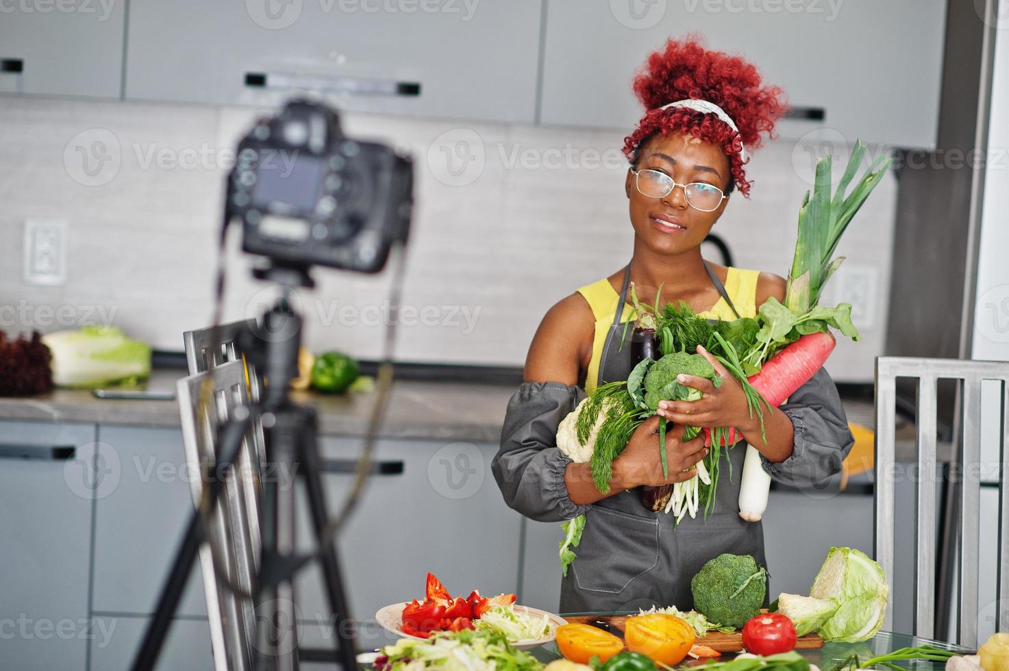 African american woman filming her blog broadcast about healthy food at home kitchen. She hold vegetables at hands. photo