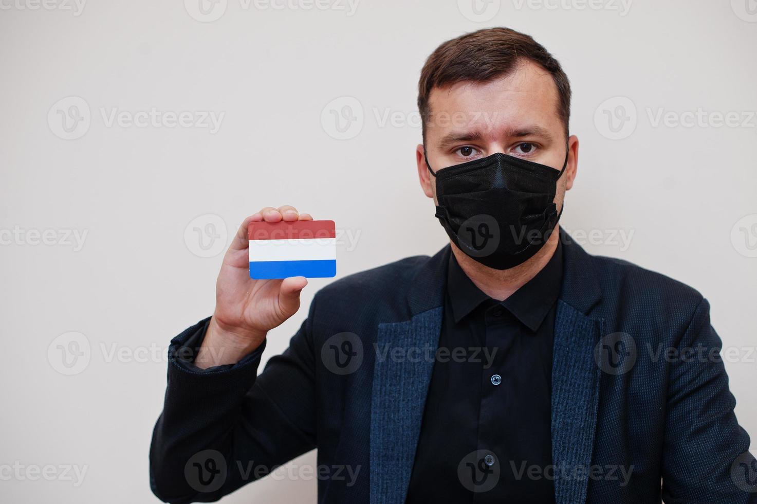 European man wear black formal and protect face mask, hold Netherlands flag card isolated on white background. Europe coronavirus Covid country concept. photo
