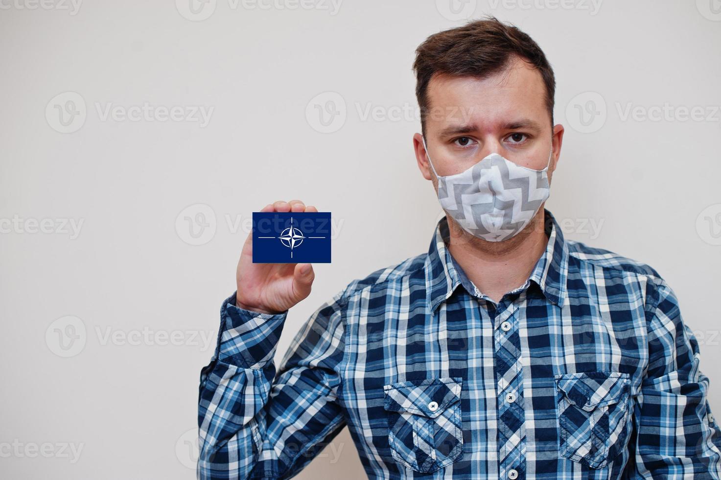 Man in checkered shirt show NATO flag card in hand, wear protect mask isolated on white background. Europe countries Coronavirus concept. photo