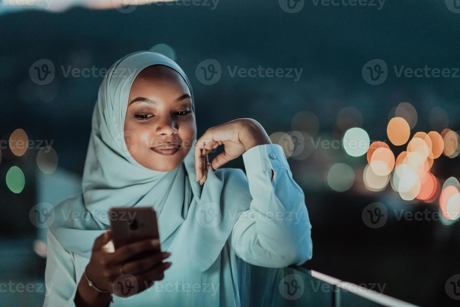 joven mujer musulmana con velo de bufanda en la calle urbana de la ciudad por la noche enviando mensajes de texto en un teléfono inteligente con la luz de la ciudad en el fondo. foto