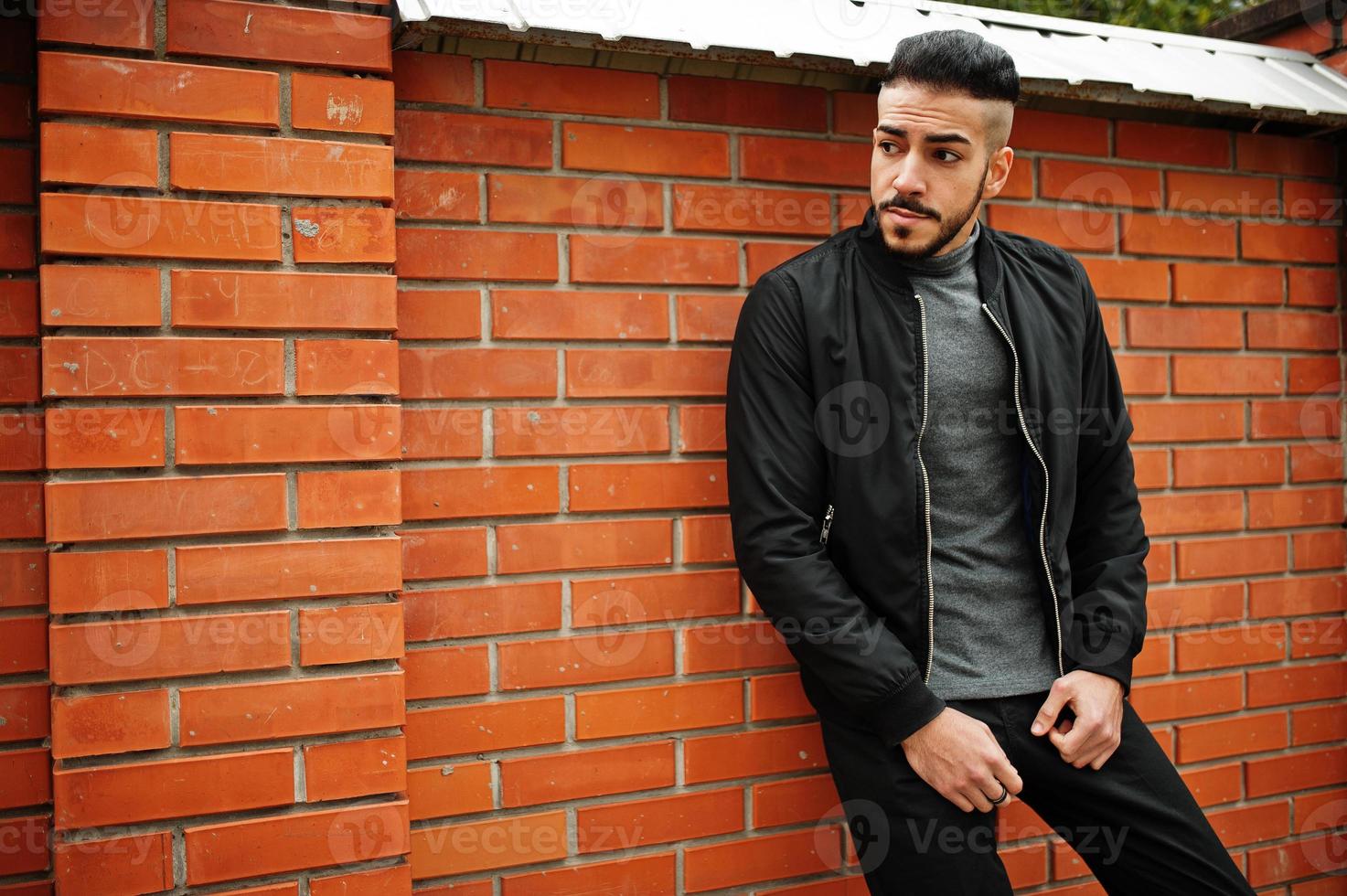 Portrait of stylish arab beard man wear grey turtleneck and black jaket. Arabian model guy against brick wall. photo