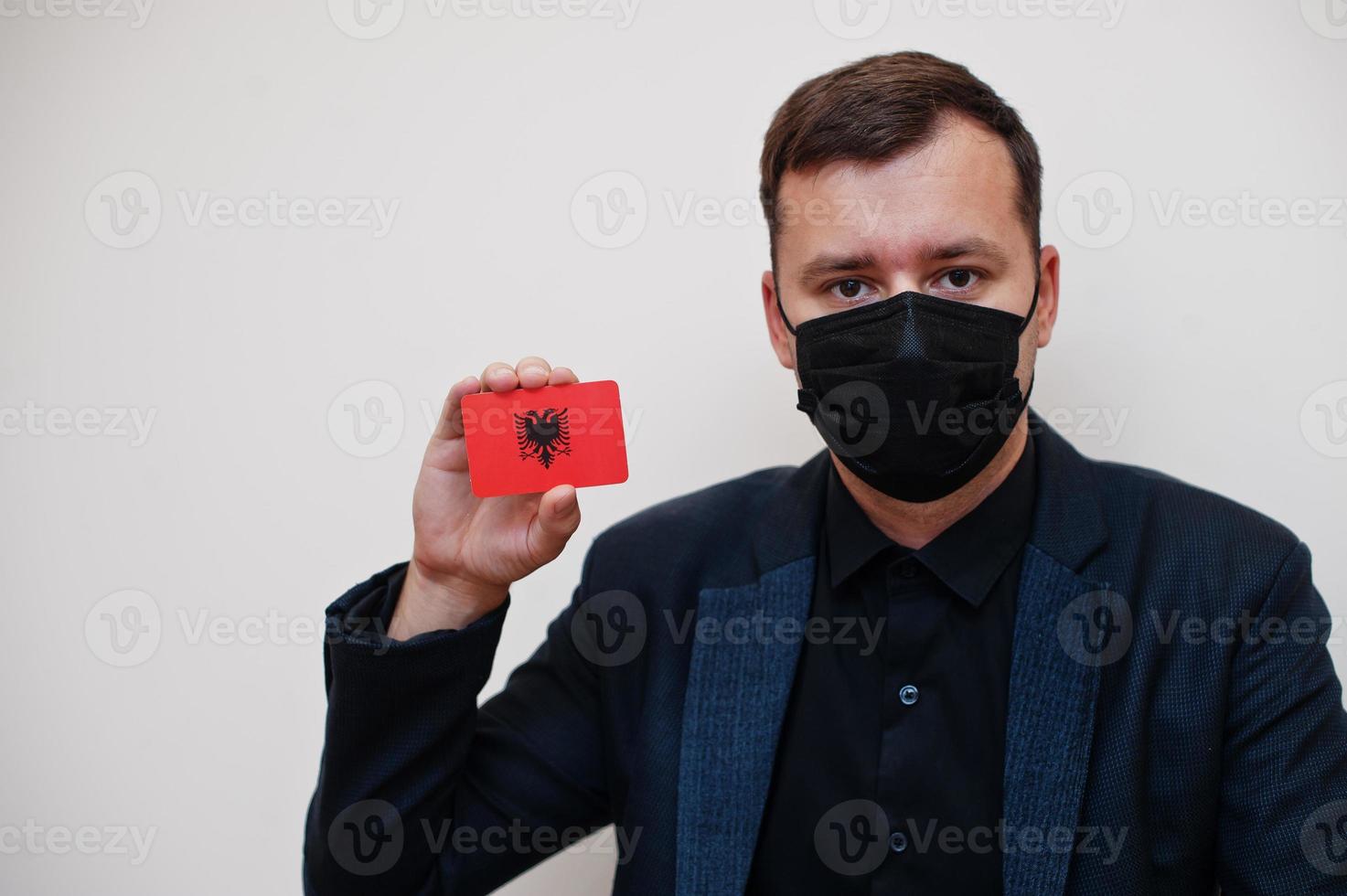 European man wear black formal and protect face mask, hold Albania flag card isolated on white background. Europe coronavirus Covid country concept. photo