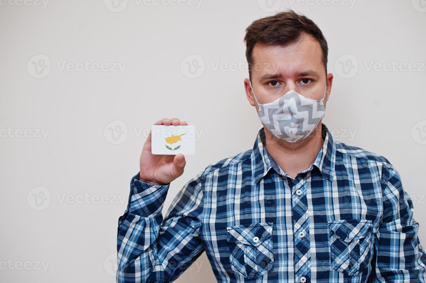 Man in checkered shirt show Cyprus flag card in hand, wear protect mask isolated on white background. Asian countries Coronavirus concept. photo