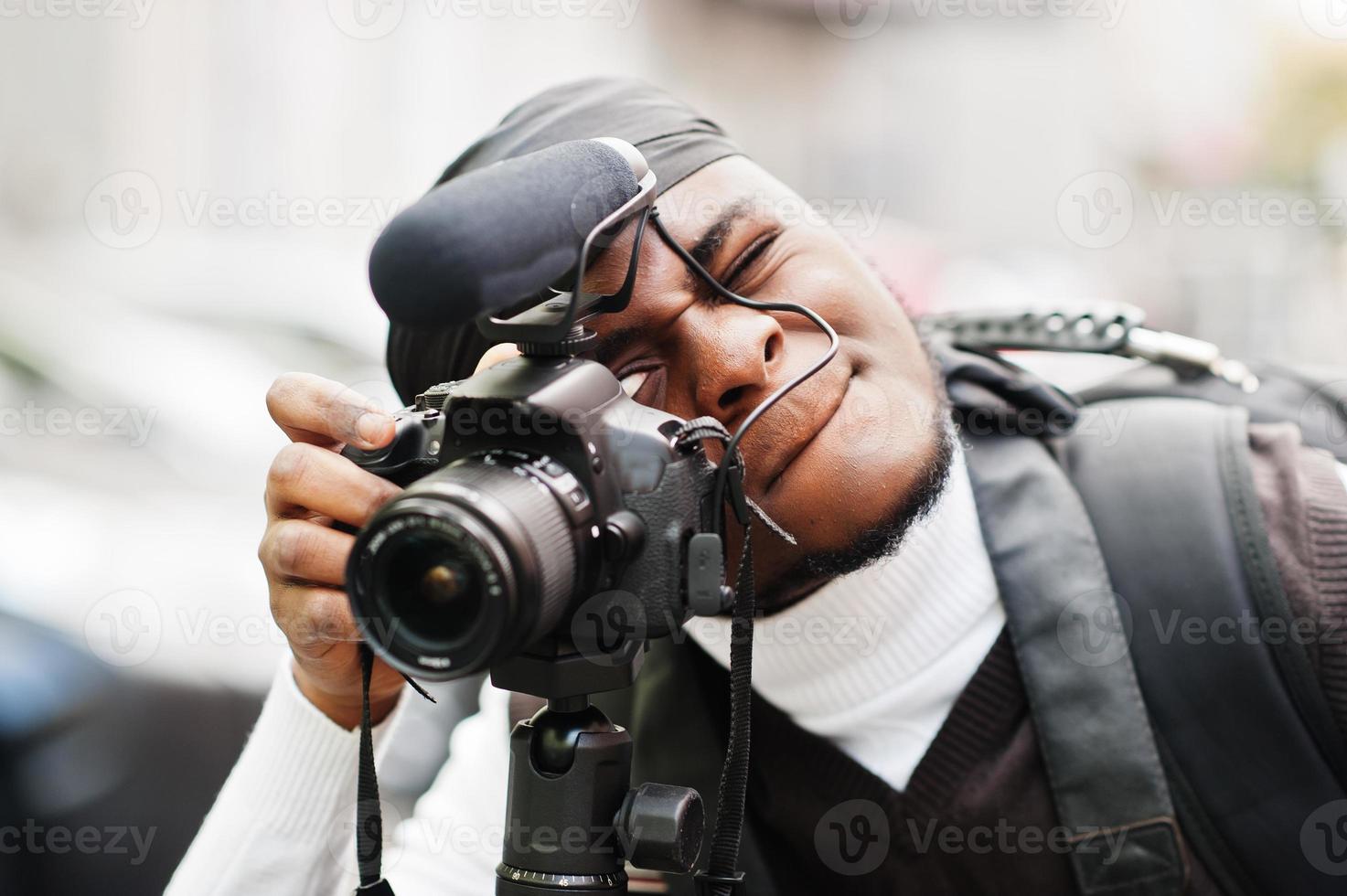 Young professional african american videographer holding professional camera with tripod pro equipment. Afro cameraman wearing black duraq making a videos. photo
