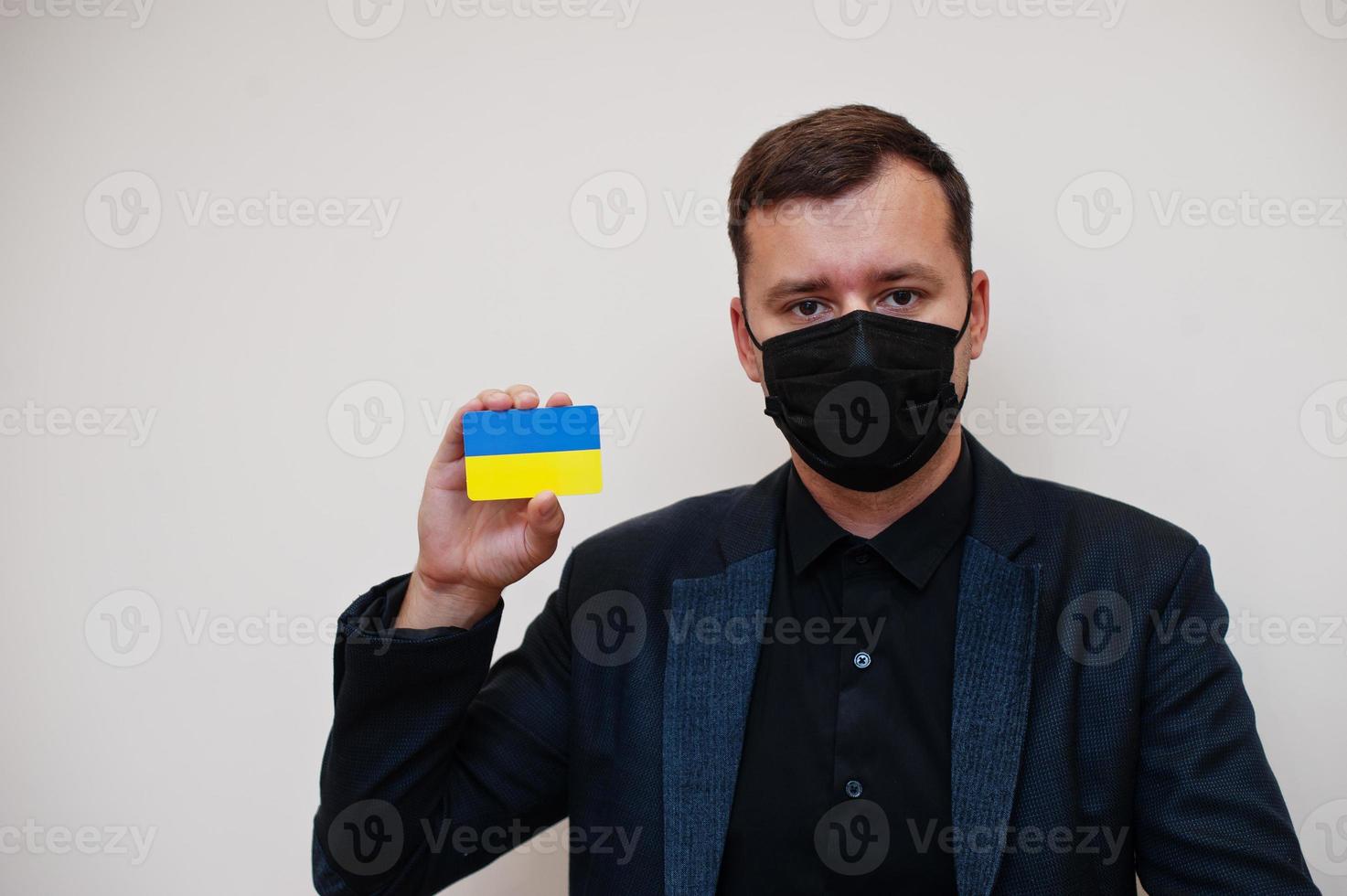 European man wear black formal and protect face mask, hold Ukraine flag card isolated on white background. Europe coronavirus Covid country concept. photo