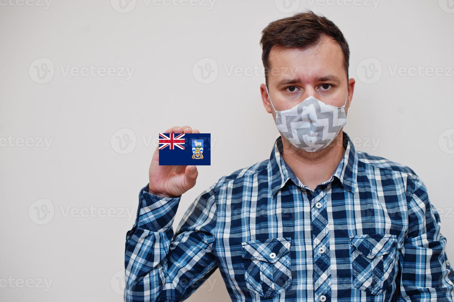 Man in checkered shirt show Falkland Islands flag card in hand, wear protect mask isolated on white background. American countries Coronavirus concept. photo