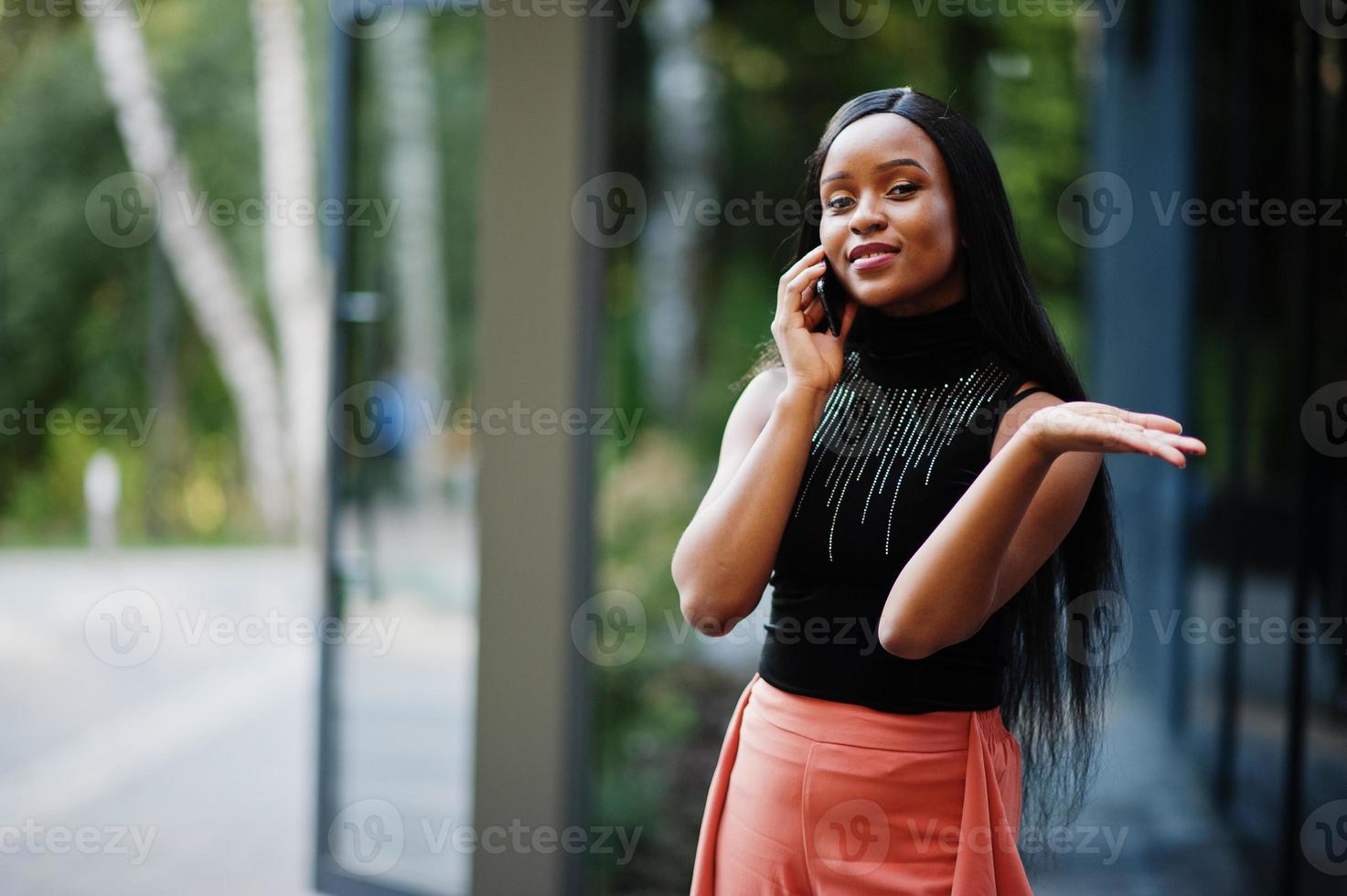 Fashionable african american woman in peach pants and black blouse speak on phone outdoor. photo