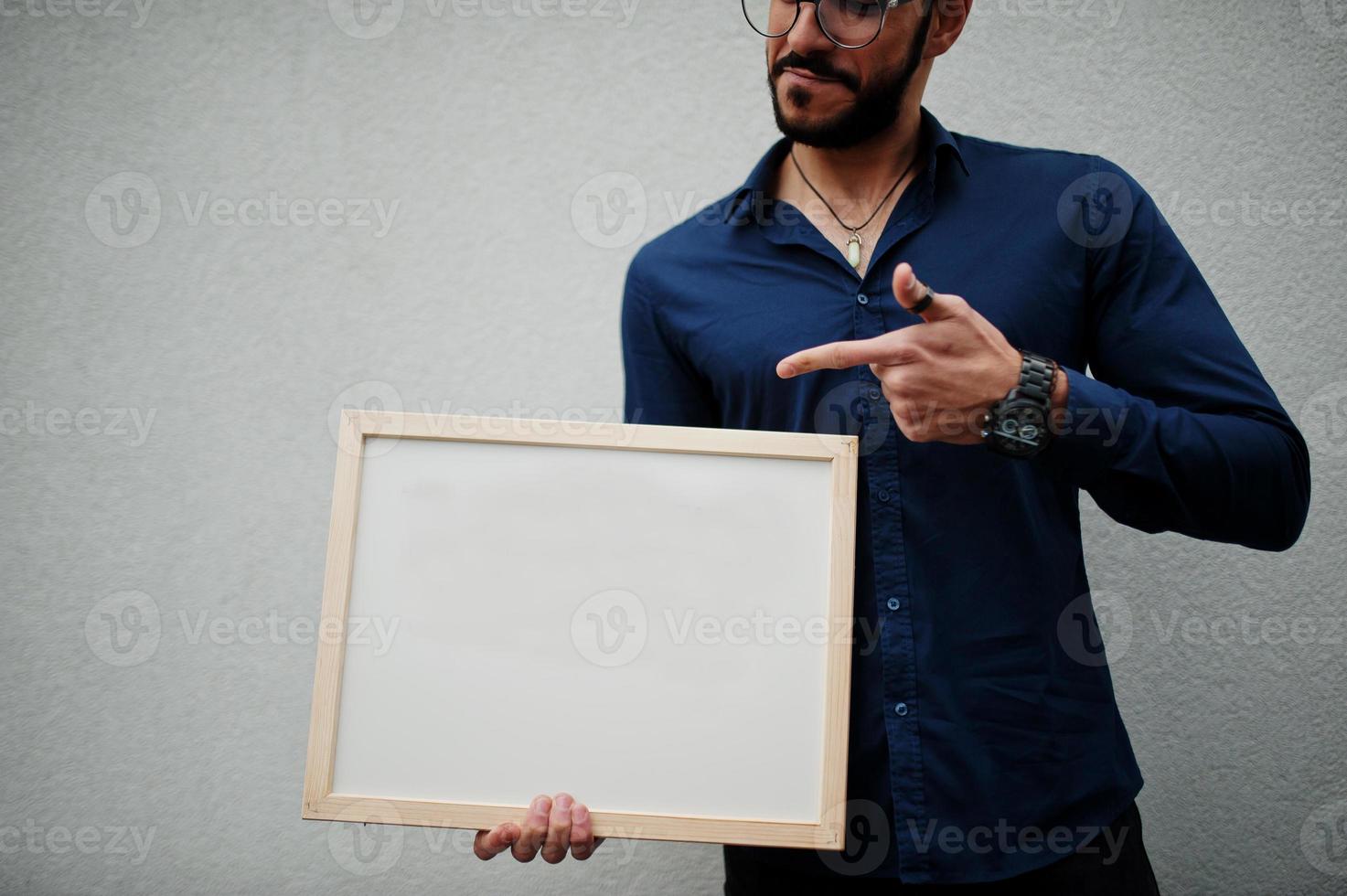 el hombre del medio oriente usa camisa azul, anteojos, sostiene un tablero blanco vacío. foto