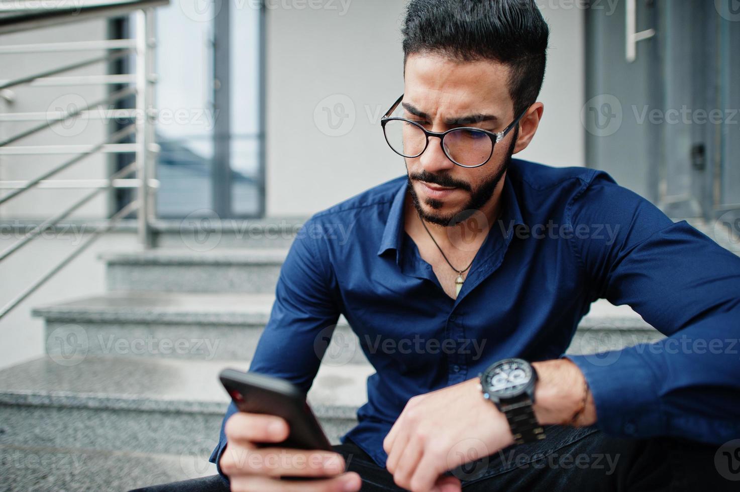 empresario del medio oriente usa camisa azul, anteojos contra el edificio de oficinas sentado en las escaleras y mira el teléfono móvil. foto