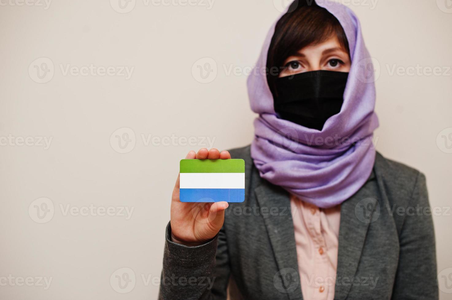 Portrait of young muslim woman wearing formal wear, protect face mask and hijab head scarf, hold Sierra Leone flag card against isolated background. Coronavirus country concept. photo