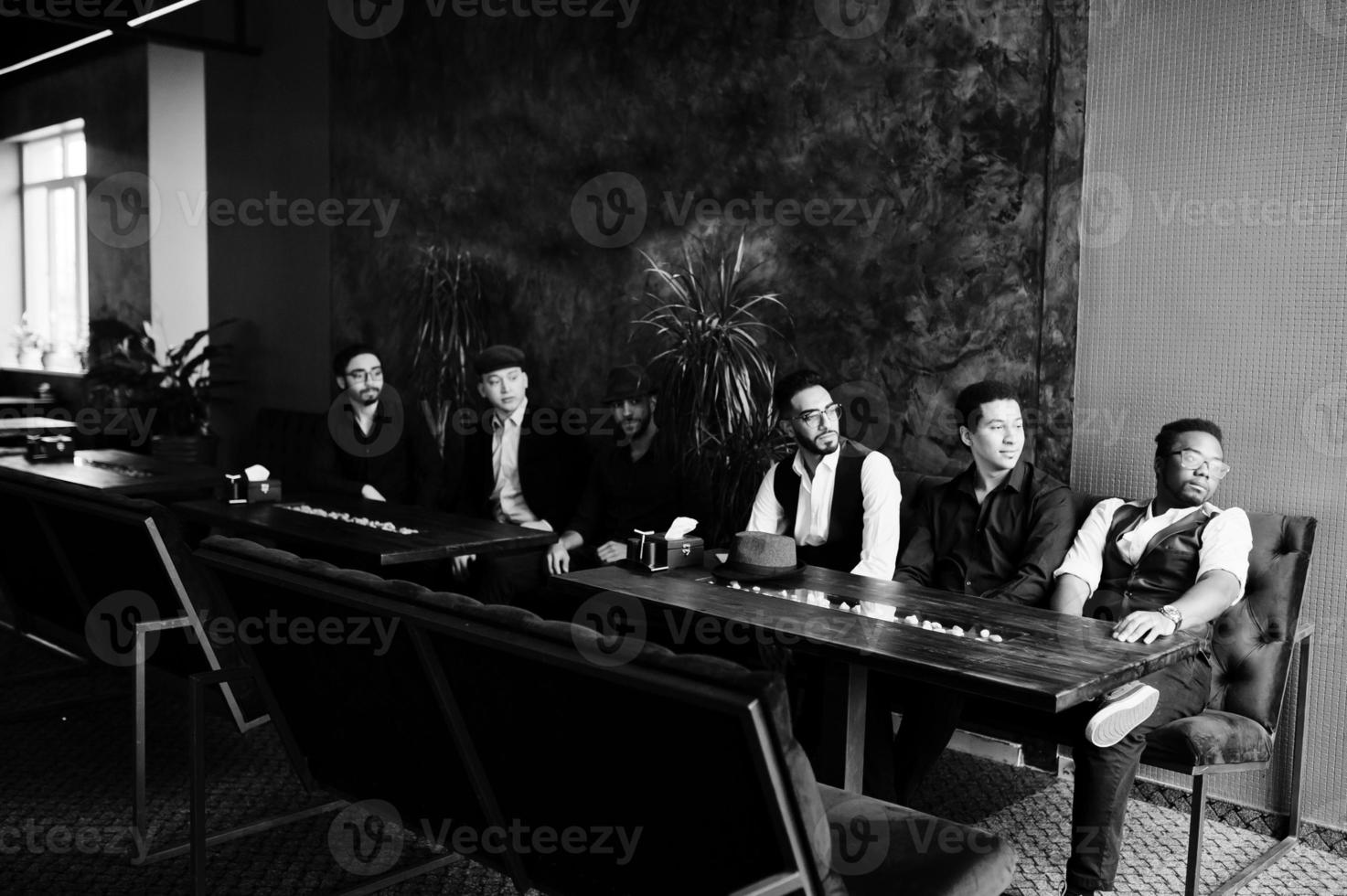 Group of handsome retro well-dressed man gangsters spend time at club. Multiethnic male bachelor mafia party in restaurant, sitting by table. photo