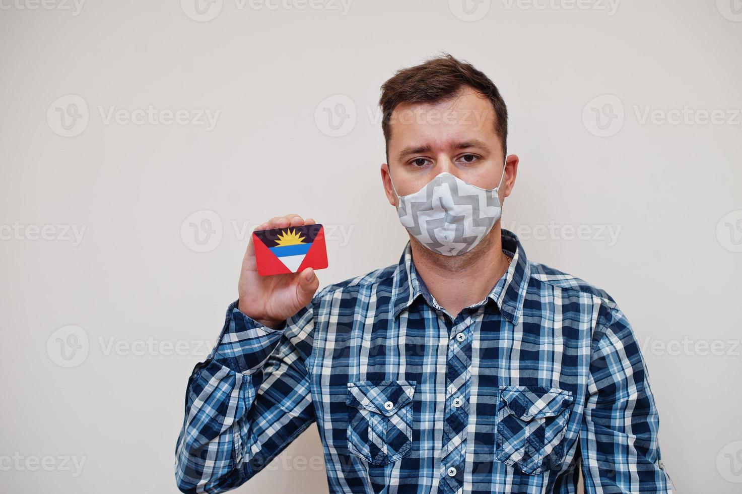 Man in checkered shirt show Antigua and Barbuda flag card in hand, wear protect mask isolated on white background. American countries Coronavirus concept. photo