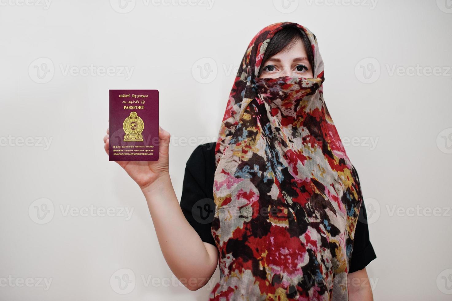 joven musulmana árabe vestida con hijab tiene pasaporte de la república socialista democrática de sri lanka sobre fondo de pared blanca, retrato de estudio. foto