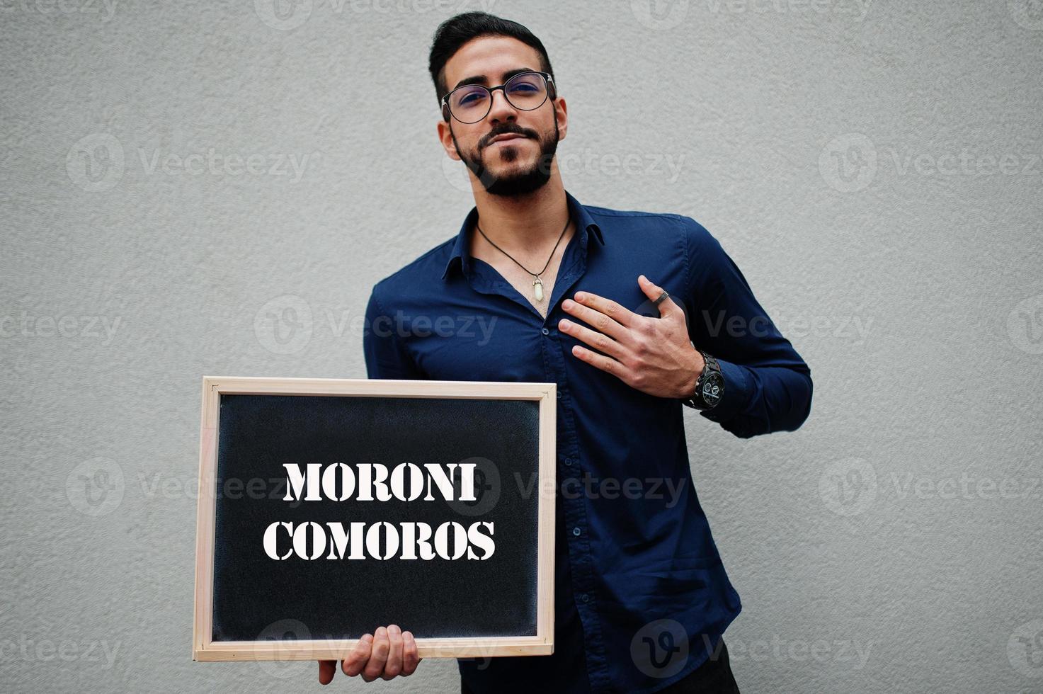Arab man wear blue shirt and eyeglasses hold board with Moroni Comoros inscription. Largest cities in islamic world concept. photo