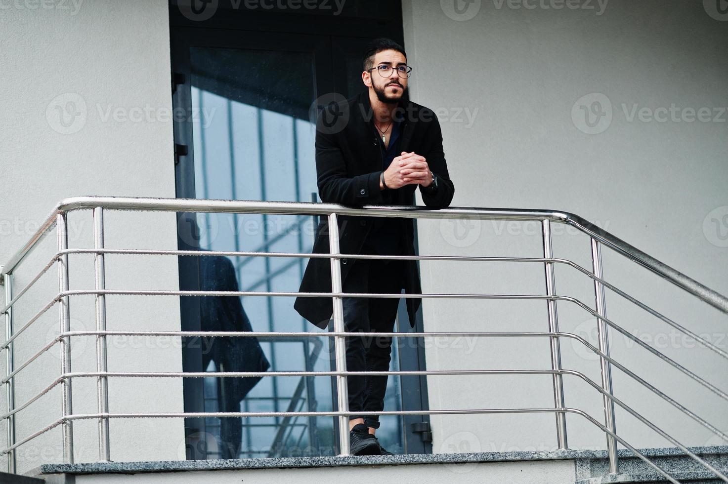 Middle eastern entrepreneur wear black coat and blue shirt, eyeglasses against office building. photo